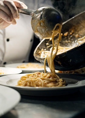 Chef pouring a serving of spaghetti with a tomato sauce from a saucepan to a plate at the pass. Photographed on location in a restaurant on the island of Møn in Denmark. Colour, horizontal with some copy space.
